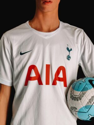 Adult soccer player in Tottenham Hotspur jersey holding a ball against a dark background.