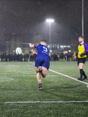 Dynamic rugby match at night in Manchester, England, played under rain and bright stadium lights.